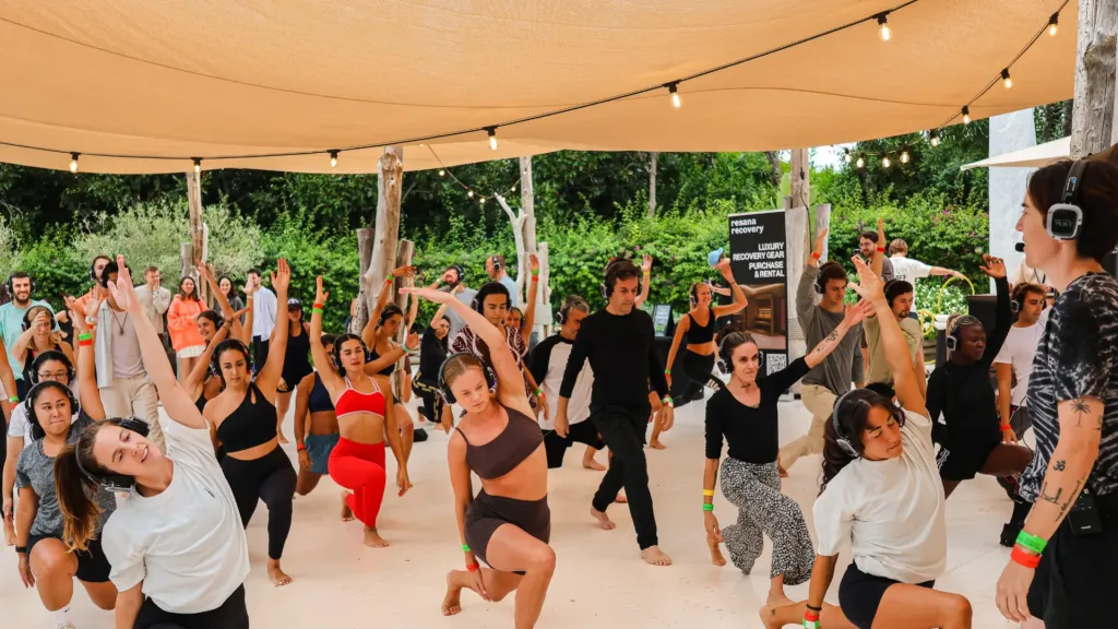 A large group in a morning movement session at a Wellness Rave event, wearing silent-disco headphones, arms raised mid-flow under a canopy outdoors