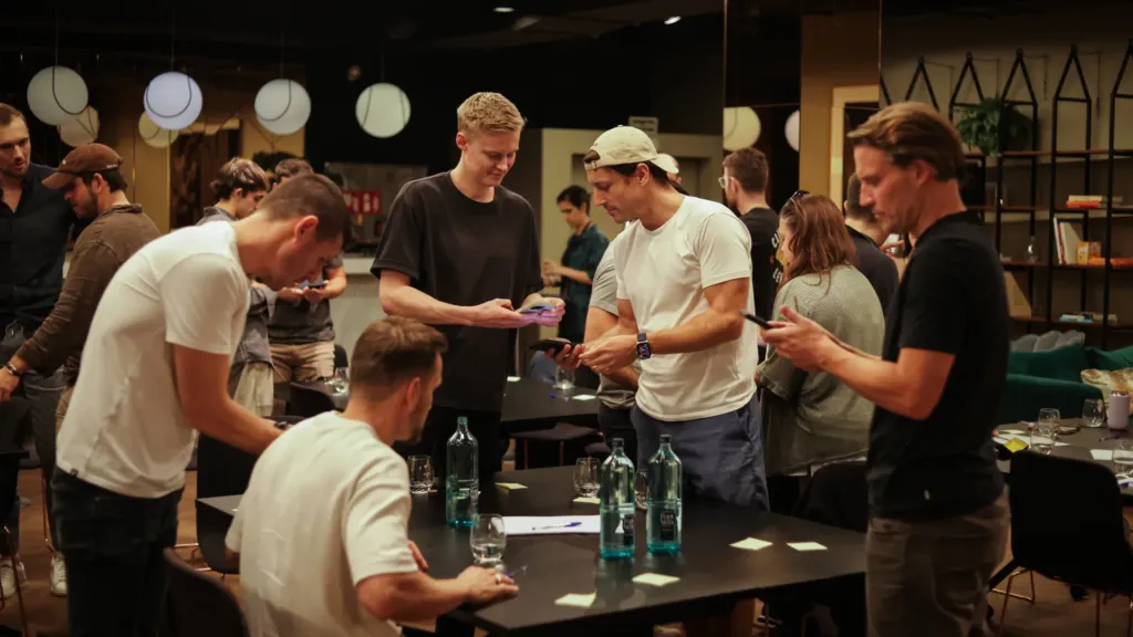 A company team gathered around tables at an in-person offsite in a modern coworking space, some talking together and some looking down at their phones.