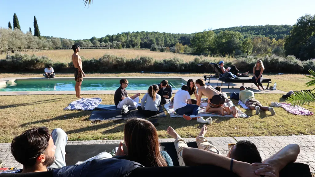 A relaxed company team gathered outdoors by a pool in the countryside, sitting on blankets and loungers in the sun